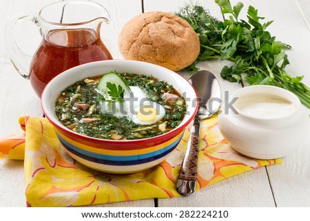Popular spring cold soup okroshka with kvass. Okroshka in a bright bowl, sour cream, kvass in a jug, a spoon, bread and herbs on white wooden table. Selective focus