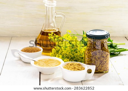 Different types of mustard: powder, seeds, cooked Dijon mustard, spicy Russian mustard, mustard oil, mustard flowers on a white wooden table. Selective focus