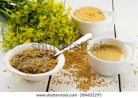 Different types of mustard: Dijon mustard and Russian, grain and mustard powder, mustard blossoms on a white wooden table. Selective focus