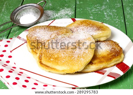 Traditional Russian fritter with powdered sugar on a plate, napkin with peas, sieve with powdered sugar on a green wooden table. Selective focus