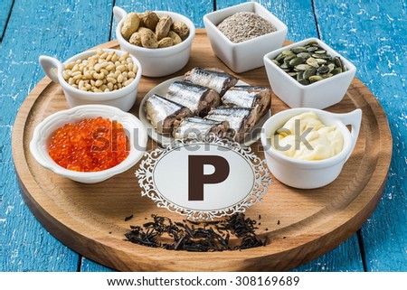 Products containing phosphorus (black tea, red caviar, pine nuts, peanuts, bran, sardines, pumpkin seeds, processed cheese) on a round cutting board and a blue wooden background