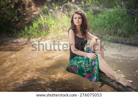 Beautiful brunette woman in bright clothes sits on the bank of the river on a background of water