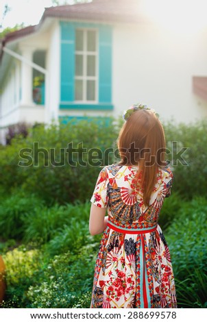 Beautiful stylish young woman in bright dress with red hair stands back and looks at the window, lifestyle, dreams, philosophy