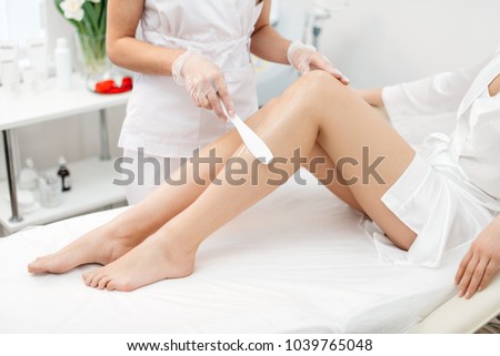 The beautician is preparing for depilation and applying the cream with wax stick on the beautiful female legs. Beauty salon.
