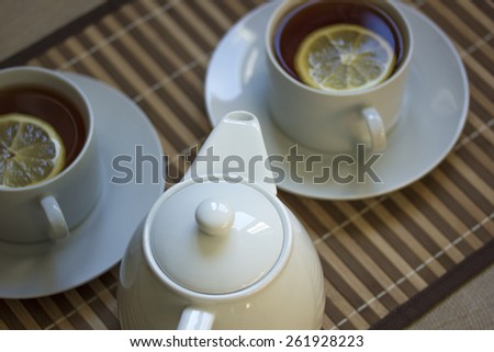 White teapot and two teacups with tea and lemon on straw plate. Shallow DOF