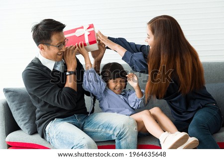 Young Asian father, mother and their cute little daughter playing with gift box on sofa at home in holiday