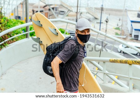 Older surfer man with covid-19 protective mask and a surfboard. Active vacation, healthy lifestyle and sports image concept