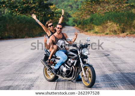 Brutal guy in blue jeans and sexy girl in shorts fun posing on a black classic motocycle on the road with trees
