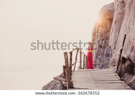Sexy girl in pink dress with a yellow belt and sunglasses posing on the pier between the rocks at sunset