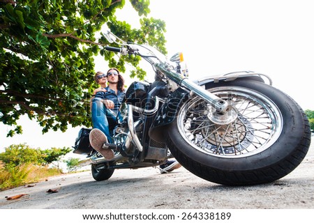 Long-haired guy in jeans and a denim shirt and sunglasses posing with a sexy girl on a motorcycle near a tree