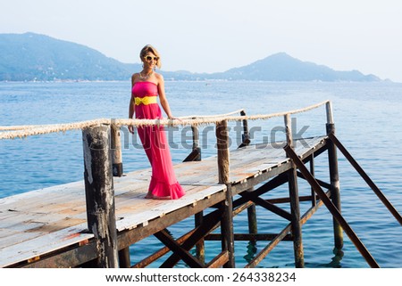 Sexy girl in pink dress with a yellow belt and sunglasses posing on the pire against the rocks at sunset