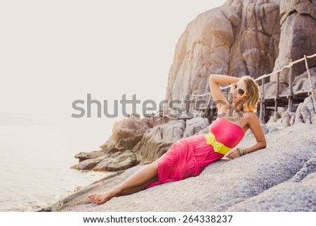 Sexy girl in pink dress with a yellow belt and sunglasses posing on the rocks at sunset