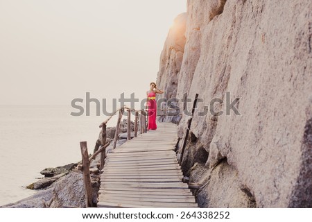Sexy girl in pink dress with a yellow belt and sunglasses posing on the pier between the rocks at sunset