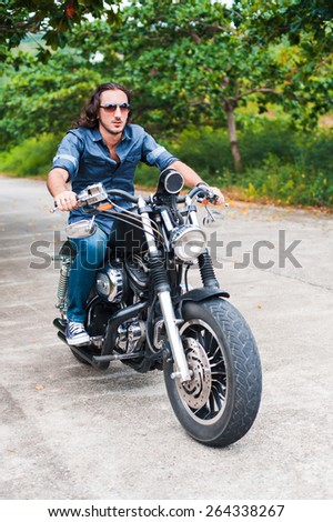 Long-haired brunette guy in sunglasses jeans and a denim shirt posing on a black ?ustom motorcycle