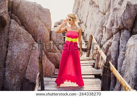 Sexy girl in pink dress with a yellow belt and sunglasses posing on the pire against the rocks at sunset