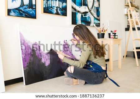 Young woman seen from behind sitting on the floor of her art studio and painting a colorful canvas with a paintbrush