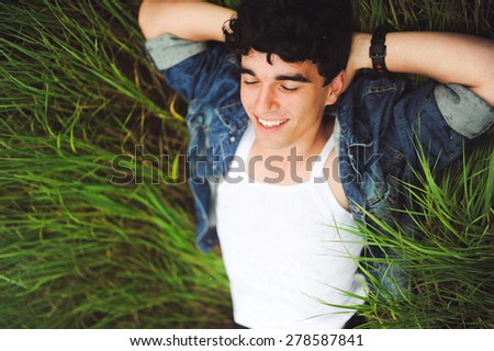 outdoor portrait of a manly  very beautiful sexy young man, dark hair, with a stunning smile, smiling and posing in the green grass in a white shirt and a denim jacket \
as the Italian