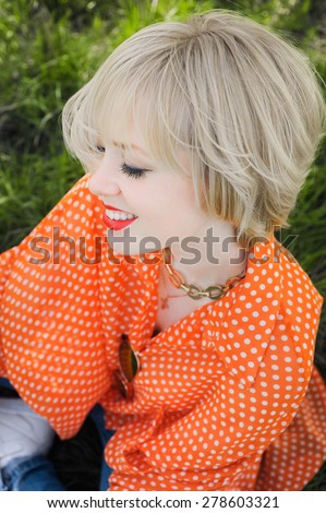 close-up portrait outdoors young beautiful girl in an orange hipster blonde bright cheerful polka dot blouse , smiling red plump lips on the background of green grass \
with white teeth