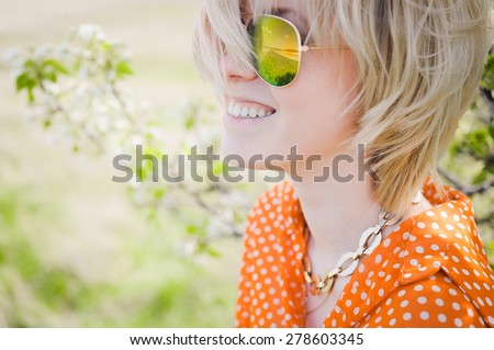 close-up portrait outdoors young beautiful girl in an orange hipster blonde bright cheerful polka dot blouse and smiling in SLR sunglasses on a background \
blooming trees  with white teeth