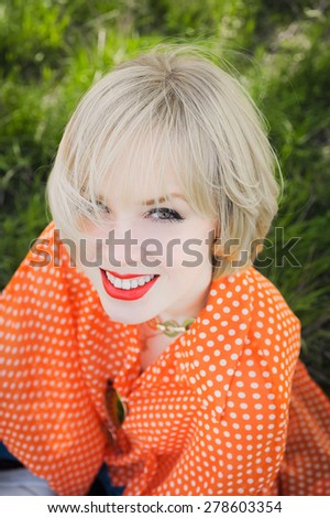 close-up portrait outdoors young beautiful girl in an orange hipster blonde bright cheerful polka dot blouse , smiling red plump lips on the background of green grass \
with white teeth