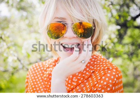 close-up portrait outdoors young beautiful girl in an orange hipster blonde bright cheerful polka dot blouse and smiling in SLR sunglasses on a background \
blooming trees  with white teeth