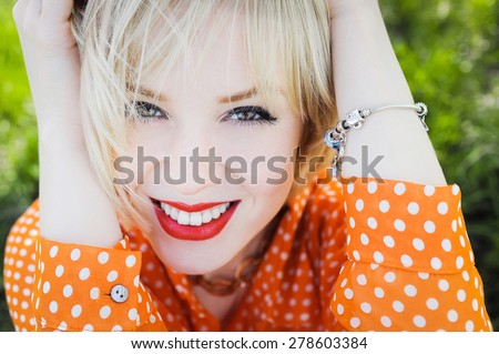 close-up portrait outdoors young beautiful girl in an orange hipster blonde bright cheerful polka dot blouse , smiling red plump lips on the background of green grass