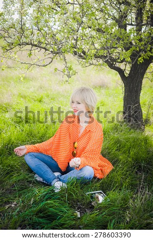 close-up portrait outdoors young beautiful girl in an orange hipster blonde bright cheerful polka dot blouse and  smiling and sitting like a yogi  on a background trees selfi yoga