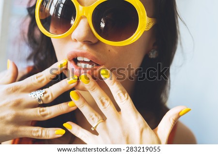 close-up portrait of a beautiful young brunette woman with full lips in yellow retro sunglasses and yellow varnish on the nails, posing and smiling for the camera
