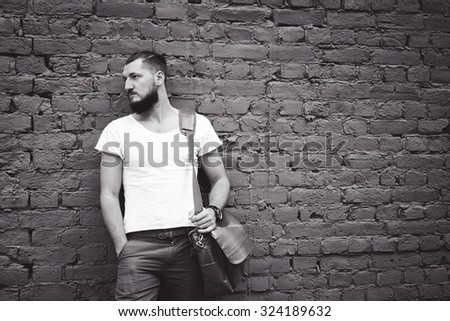 young guy with a beard and mustache  and white T-shirt  posing on the street  vintage man, fashion men, hipster street casual  leather bag and hours against the background of a brick wall