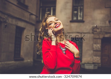 stylish portrait of a beautiful young blonde with red lips and curly hair talking on the phone on the background of the street smiling and posing in a red blouse