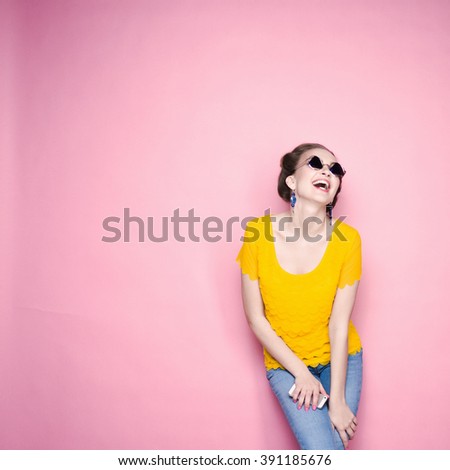 close-up portrait of a beautiful young blonde girl in fashionable sunglasses on a pink background in the studio in a yellow blouse and jeans holding a popular phone smiling