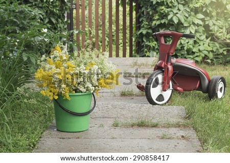 Child\'s bicycle on a sidewalk with a green bucket of white and yellow flowers. Summer morning in a village house