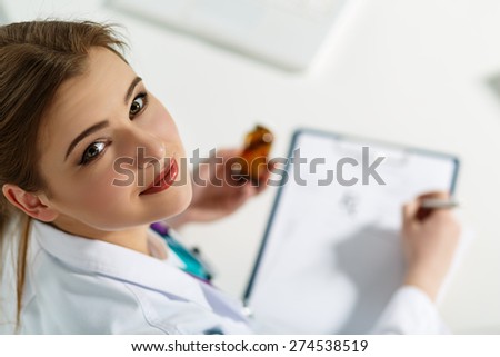 Beautiful young doctor sitting in front of working table , holding container with pills and writing prescription. Medical concept. Handsome female doctor distracted from paperwork looking up