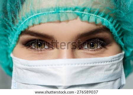 Female doctor\'s face wearing protective mask and green surgeon\'s cap closeup. Surgeon\'s eyes close up gazing intently in camera. Resuscitation concept.