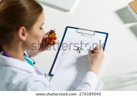 Beautiful young doctor sitting in front of working table, holding container with pills and writing prescription. Medical or pharmacy concept. Handsome female doctor doing paperwork with concentration