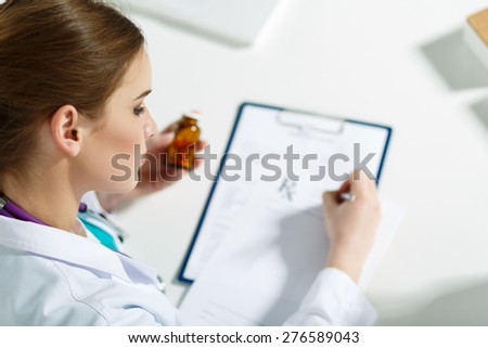 Beautiful young doctor sitting in front of working table, holding container with pills and writing prescription. Medical or pharmacy concept. Handsome female doctor doing paperwork with concentration