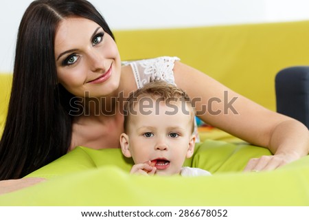 Mother and son playing and looking in camera. Beautiful young smiling woman sitting behind her little boy. Childhood and parenthood concept. Baby-sitter with kid