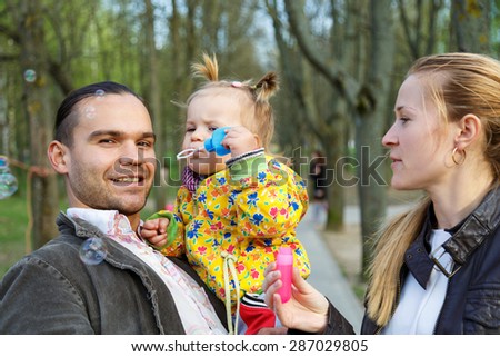 Happy parents with daughter outdoor playing and making soap bubbles. Beautiful smiling woman and handsome man blowing bubbles to cute little girl in city park. Childhood and parenthood concept