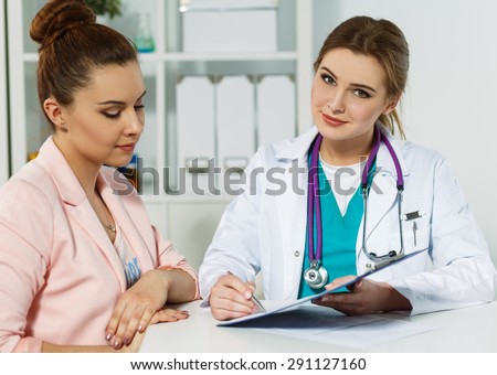 Beautiful smiling female medicine doctor holding clipped pad with document and pointing in it with pen to patient. Signing treatment bill or insurance policy concept