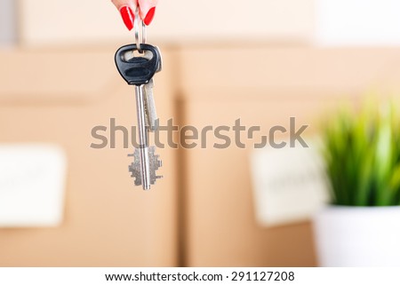 Female hand holding keys over pile of brown cardboard boxes with house or office goods background. Moving to new place of living concept.