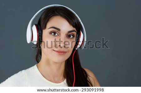 Beautiful dark haired smiling woman wearing headphones, holding phone and listening music on grey background. Modern urban life concept