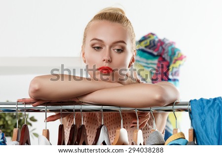 Thoughtful sad beautiful blonde woman standing near wardrobe rack full of clothes and choosing dress. Shopping and consumerism or stylist concept. Nothing to wear and hard to decide concept