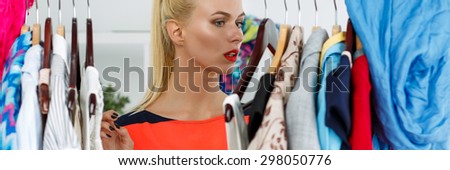 Thoughtful sad beautiful blonde woman standing near wardrobe rack full of clothes and choosing dress. Shopping and consumerism concept. Nothing to wear and hard to decide concept. Letterbox view