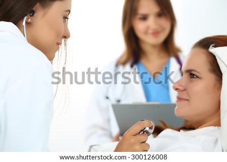 Female medicine doctor communicating, examining and listening with stethoscope patient during ward round while nurse filling in patient medical history list in background. Focus on phonendoscope head