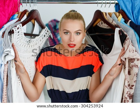 Beautiful smiling blonde woman standing inside wardrobe rack full of clothes suffering with choice. Shopping and consumerism or stylist concept. Nothing to wear and hard to decide concept