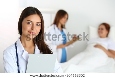 Beautiful female medicine doctor looking in camera in front of patient lying in bed and communicating with therapeutist. Physician or traumatologist medical concept. Medical care or insurance concept