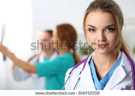 Beautiful smiling female medicine doctor looking in camera portrait while two colleagues examining x-ray picture in background. Radiology, traumatology, medical care or insurance concept