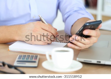 Male hand holding silver pen ready to make note in opened notebook while looking at cellphone. Businessman or employee at workplace writing business ideas, plans or tasks at personal organizer