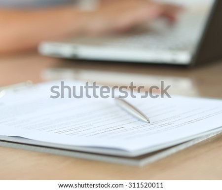 Silver pen lying on document pad while female hands working on notebook pc in background. Office life, paperwork, client contract, business agreement concept. Focus on pen