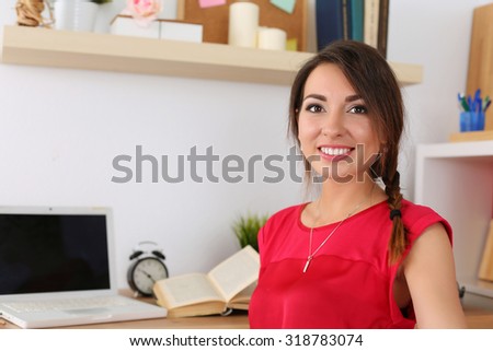 Beautiful smiling female student sitting in her room half turn studying. Young brunette woman in red dress at workplace with laptop and opened book preparing for exams. Education and self development